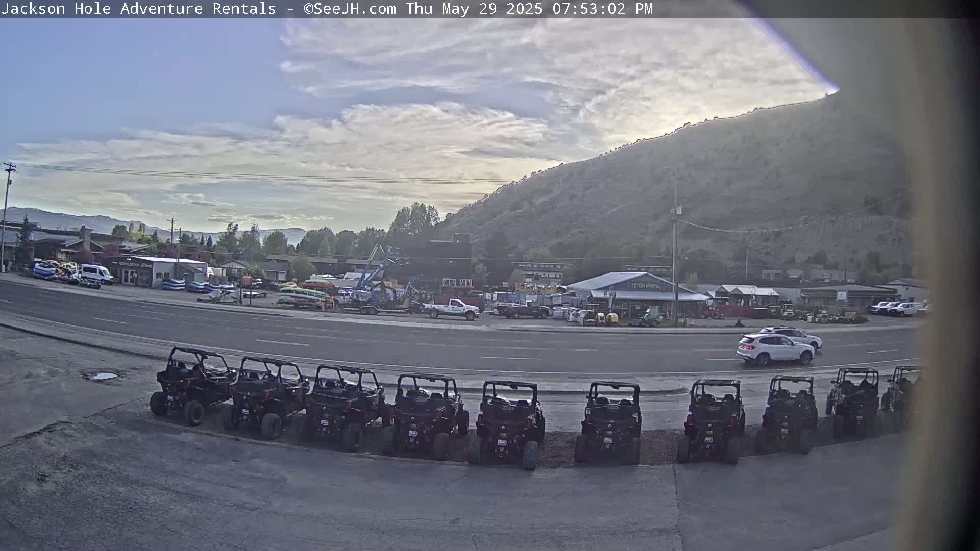 A row of parked all-terrain vehicles sits in a parking lot next to a road, with a variety of buildings and a large hill in the background under a partly cloudy sky.