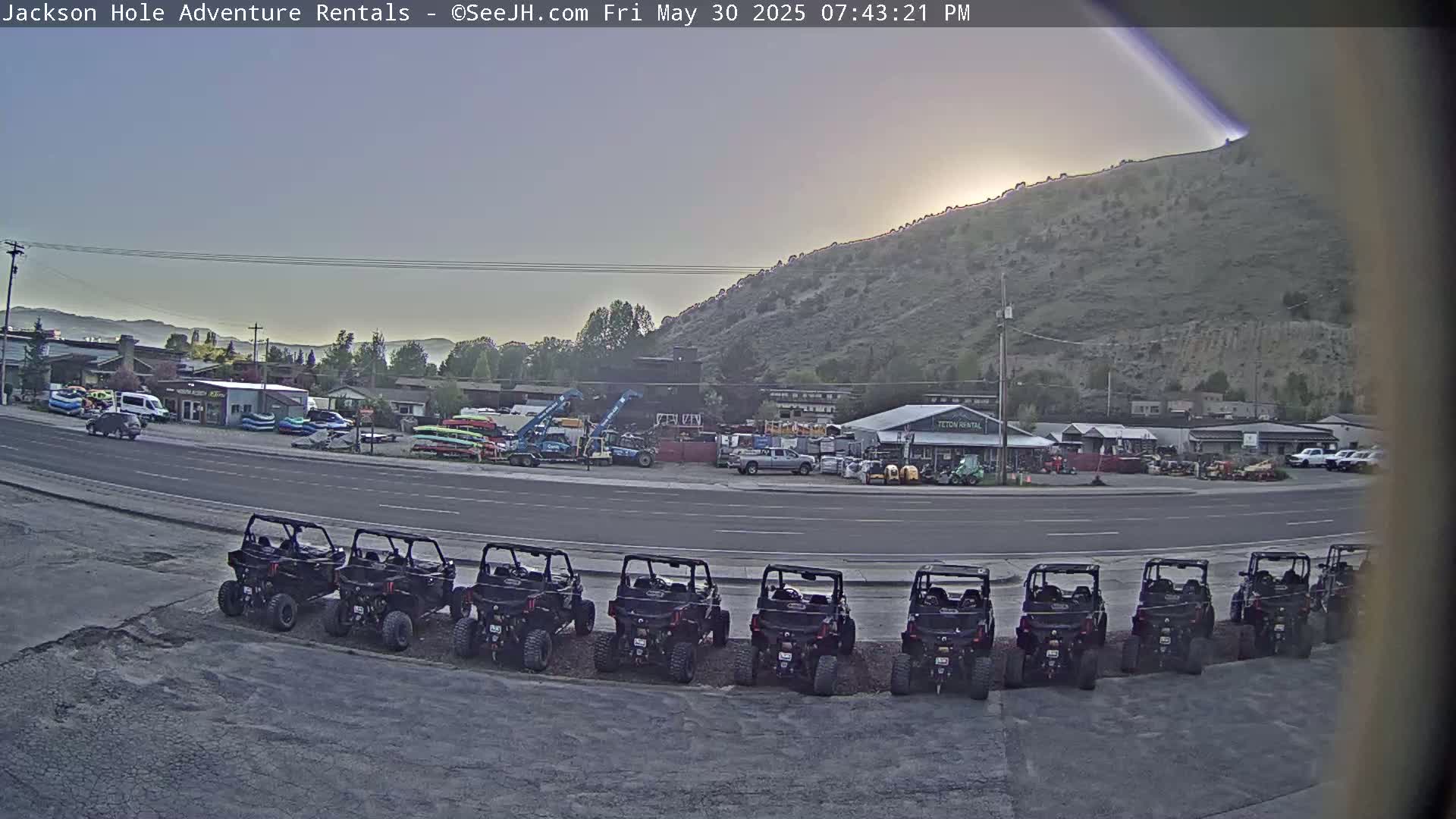 A row of all-terrain vehicles is parked in a lot next to a road, with a hill and various buildings in the background under a partly sunny sky.