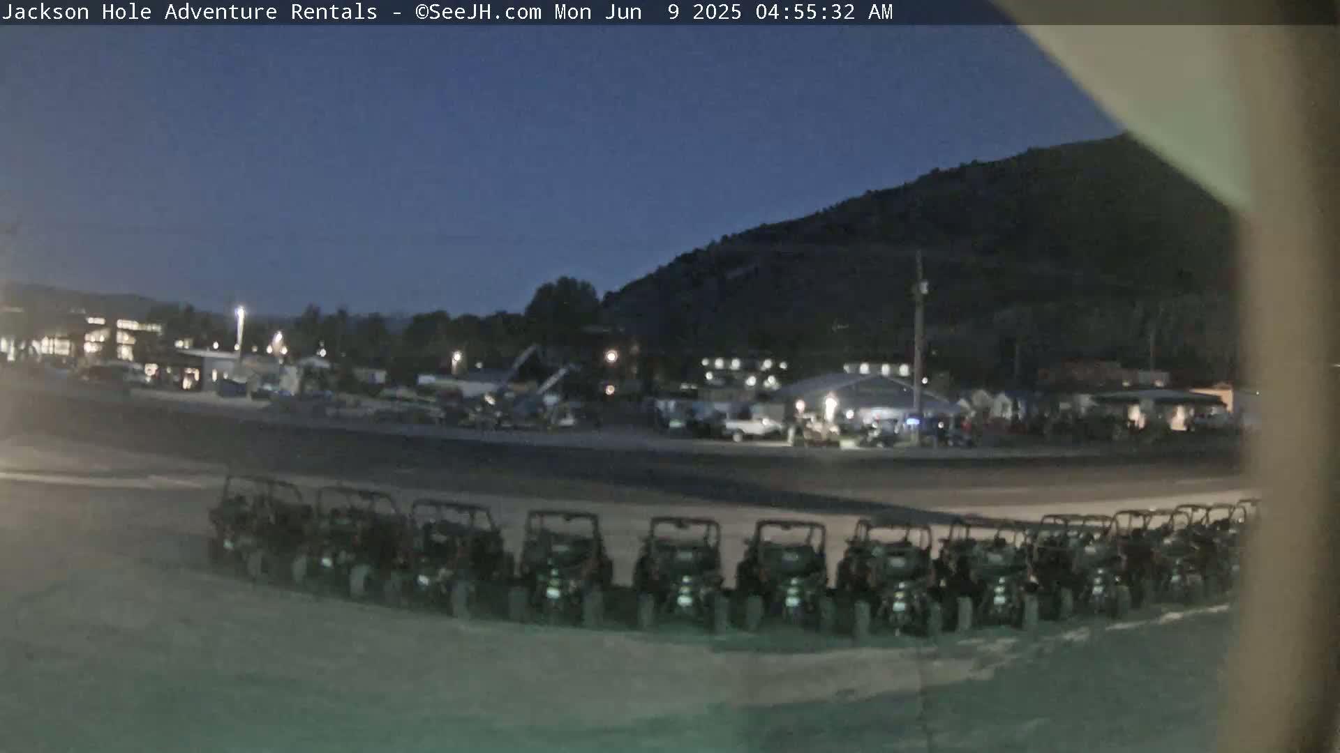 A row of all-terrain vehicles is parked in a lot at dusk, with a dimly lit town and a dark mountain in the background.