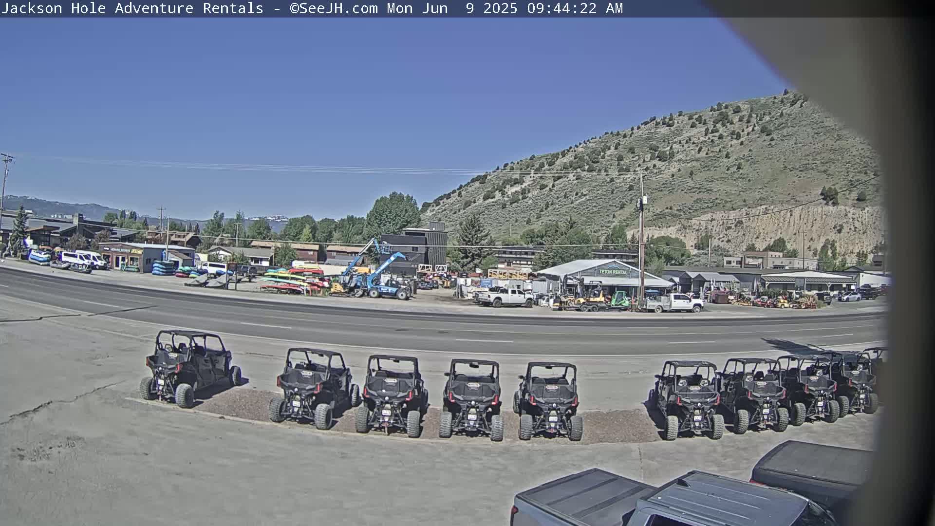 Under a clear blue sky, several off-road vehicles are parked in a lot next to a roadway, in front of a business that rents outdoor recreational equipment.