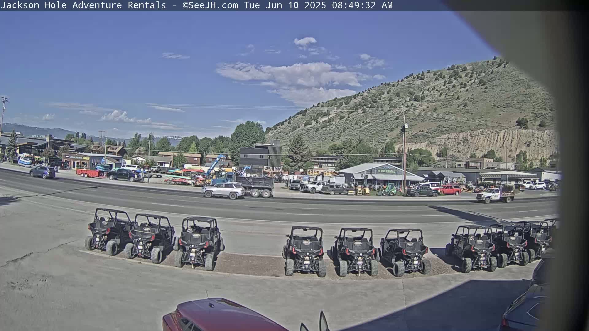 Under a mostly sunny sky, a row of all-terrain vehicles is parked in front of a building complex, across the street from a variety of other vehicles and equipment.