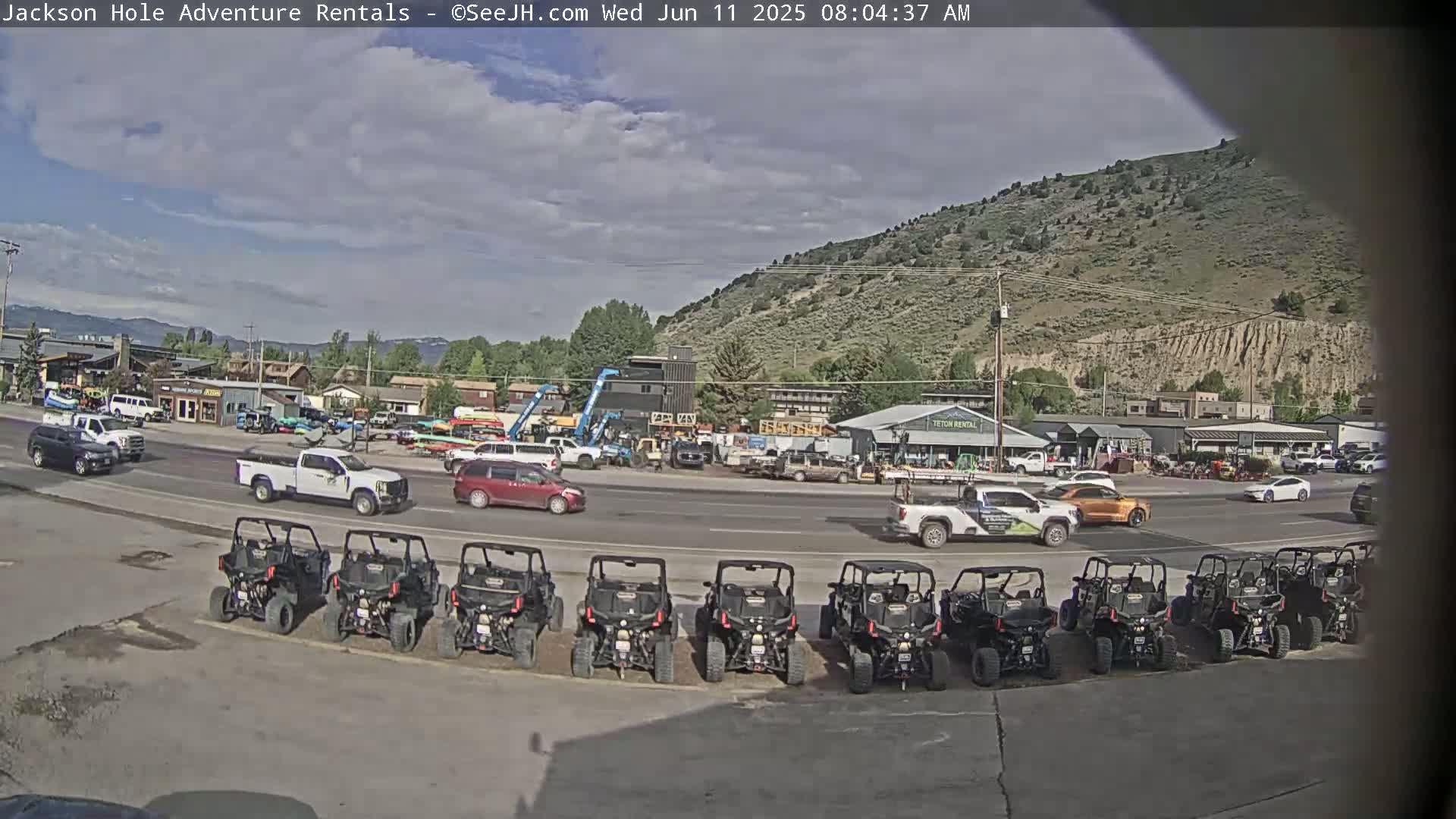 A mostly sunny day shows a row of parked all-terrain vehicles in front of a street with several cars and trucks, all near a collection of businesses and a hillside in the background.