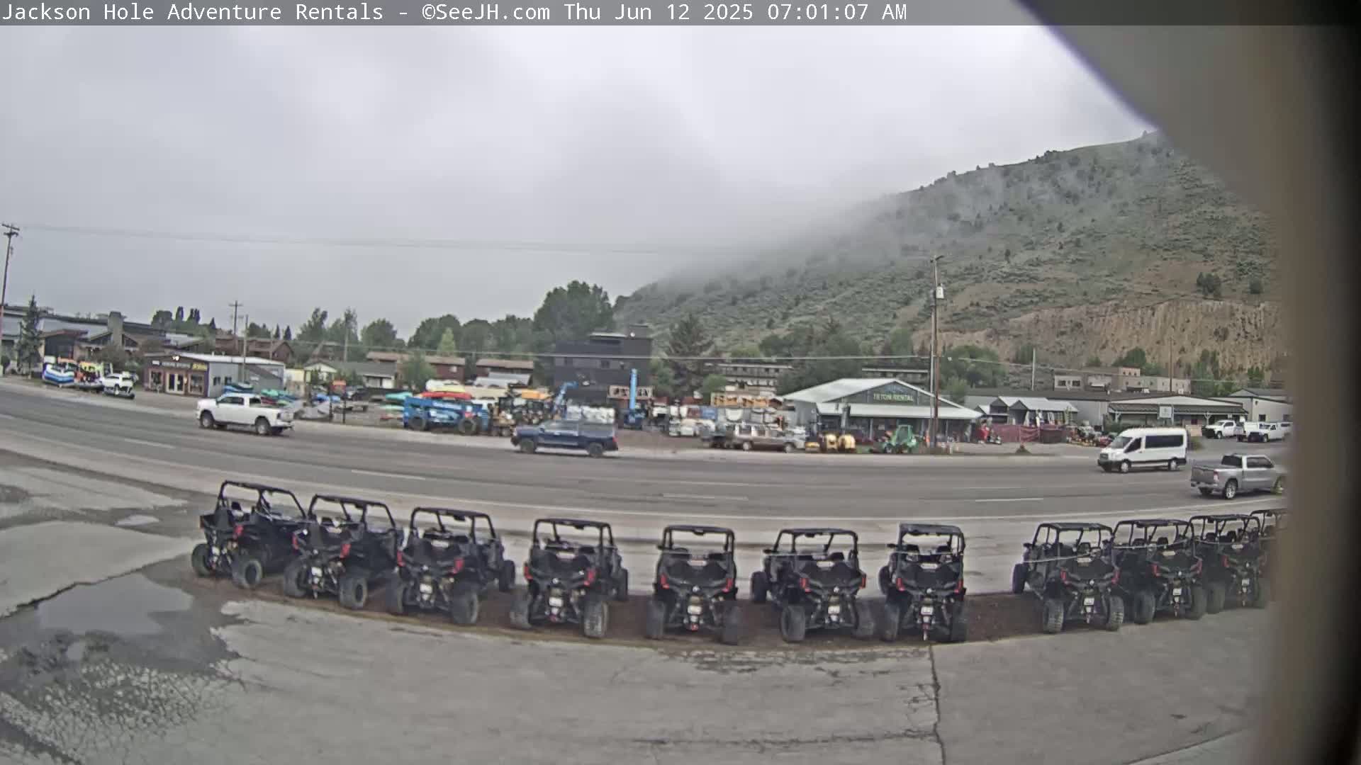 A row of off-road vehicles is parked in a lot next to a road, in front of several buildings and a hill shrouded in fog.