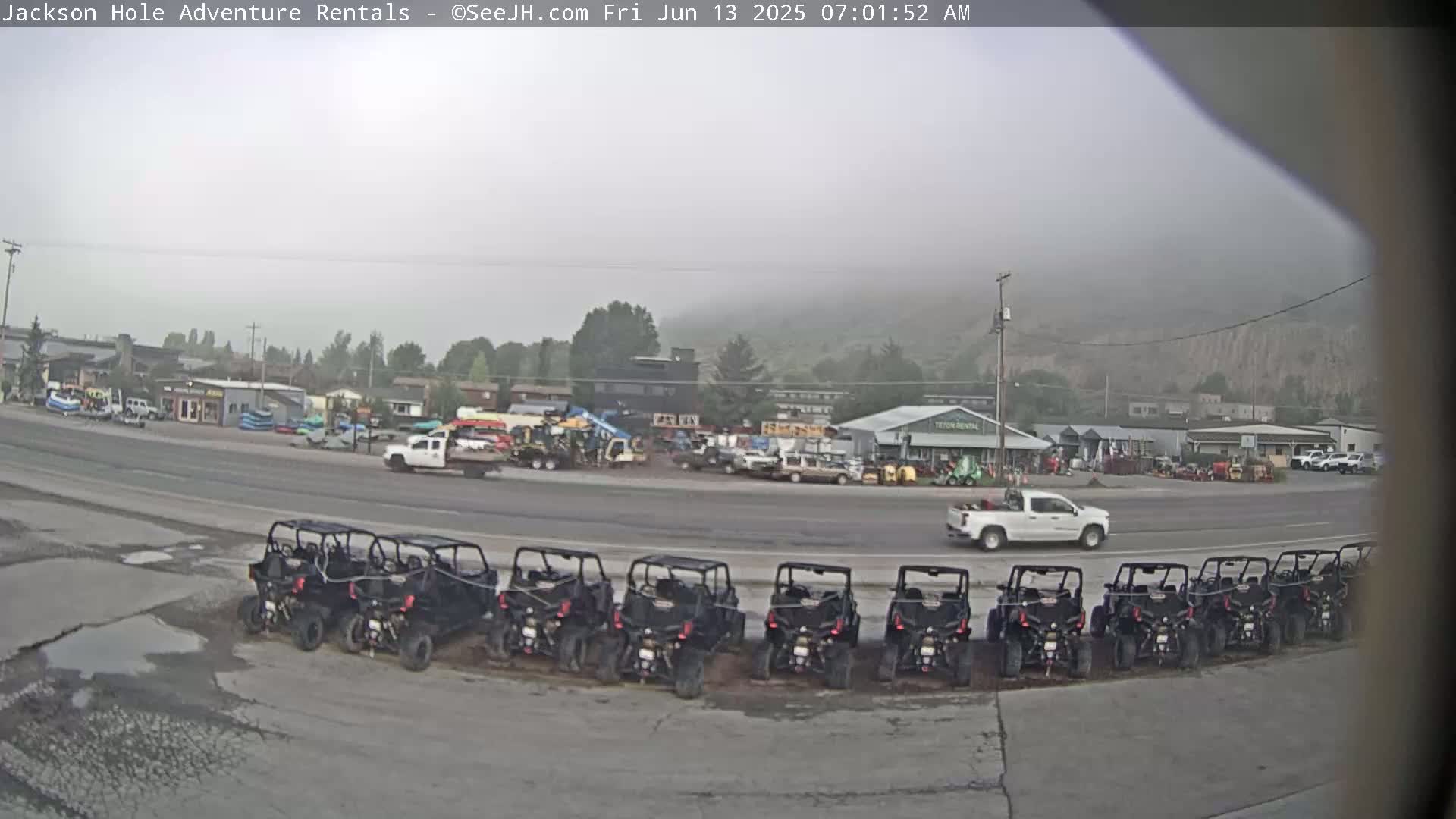 A row of all-terrain vehicles is parked in a lot next to a street with several buildings and vehicles visible in the hazy, overcast morning.