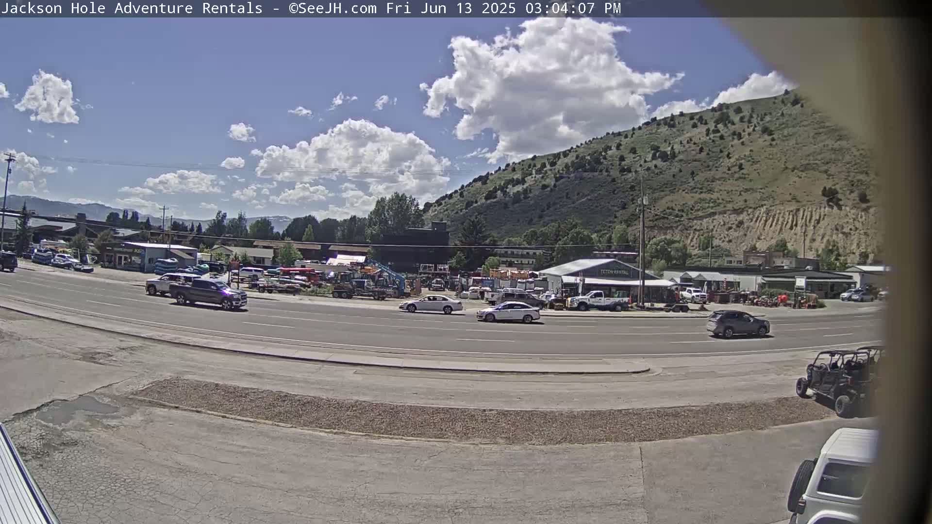 A partly sunny day reveals a road scene with several vehicles, businesses, and a large, green hill in the background.