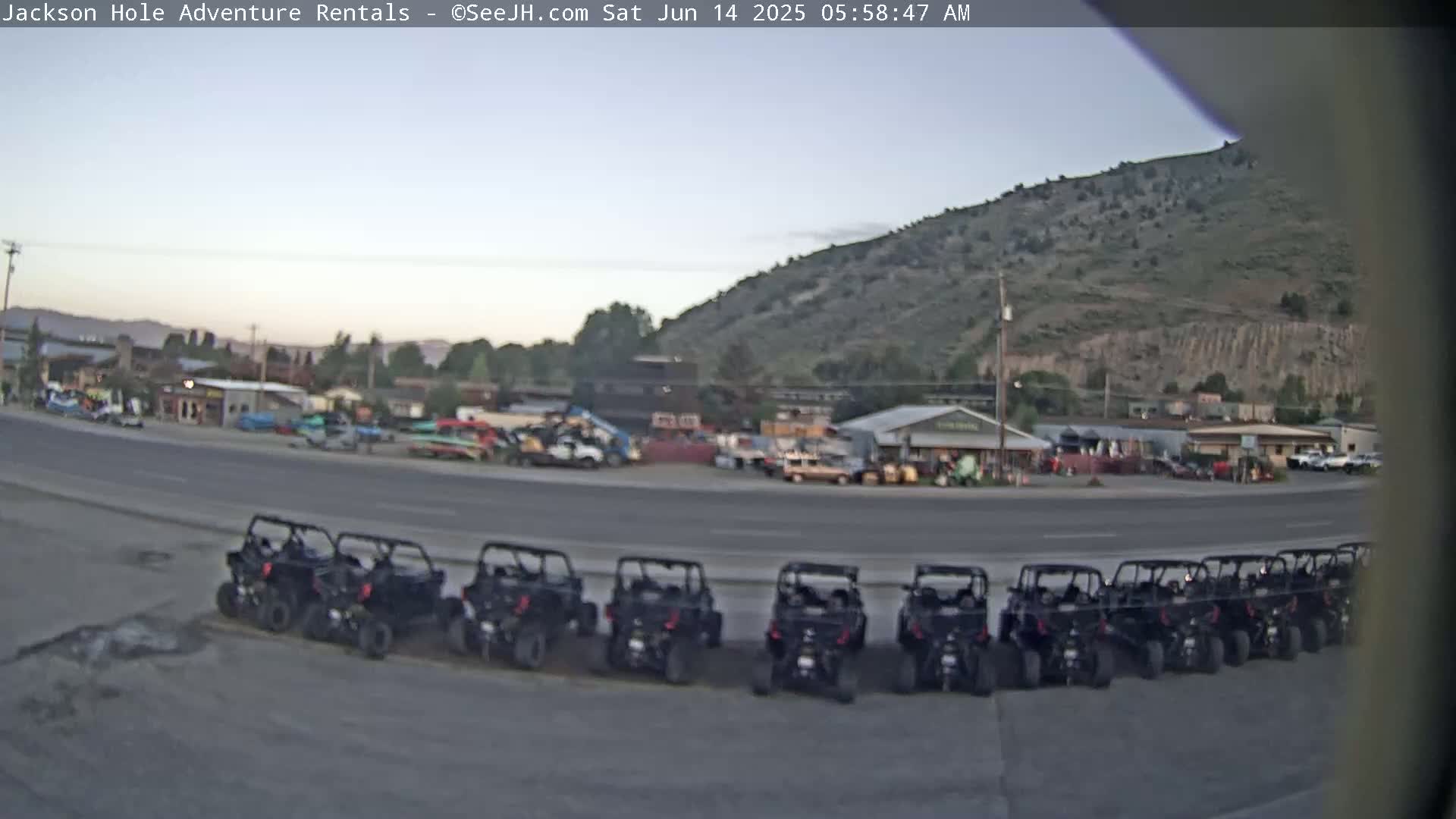 A row of all-terrain vehicles is parked in a lot next to a road in front of several buildings, under a partly cloudy sky.