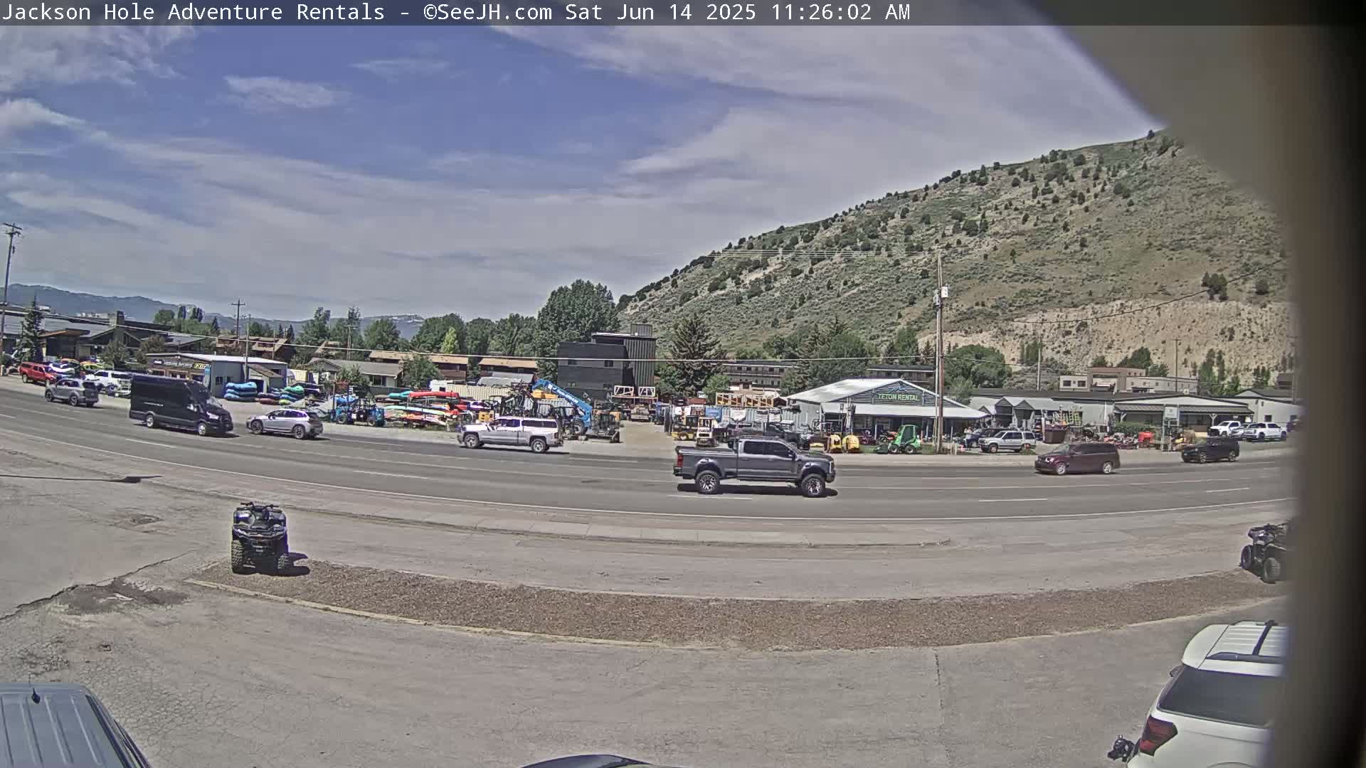 A sunny day reveals a street scene with several vehicles, including trucks and an ATV, parked near a row of businesses and a background of a hill.