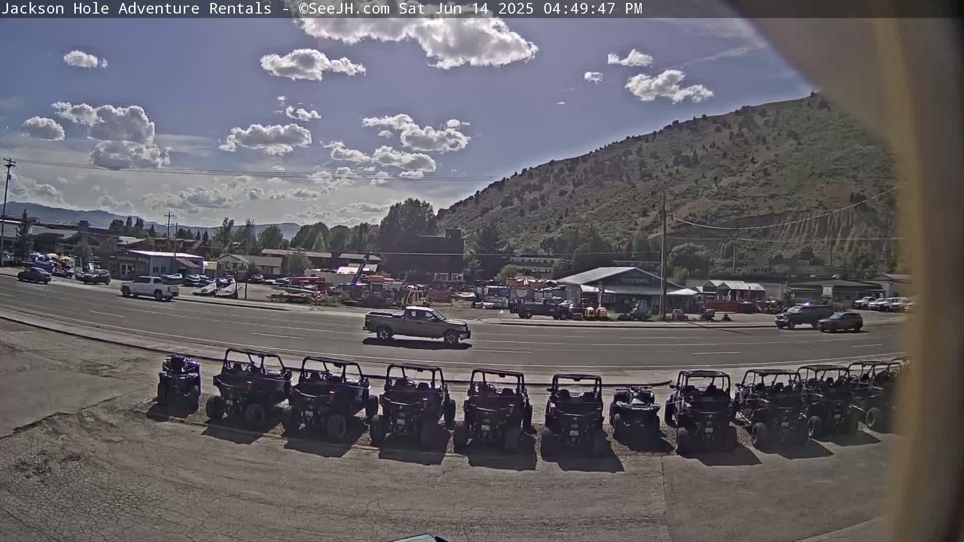 A sunny day reveals a street scene with several vehicles, including trucks and an ATV, parked near a row of businesses and a background of a hill.