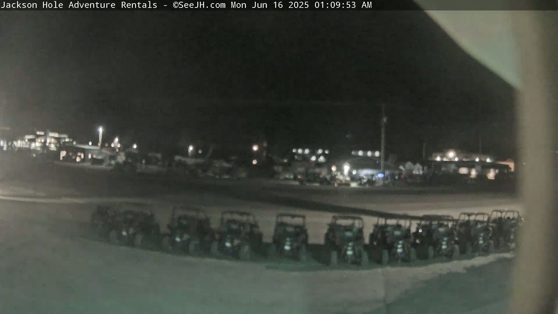 A nighttime, outdoor scene shows a row of parked all-terrain vehicles in a parking lot, with a dimly lit building and other vehicles visible in the background.
