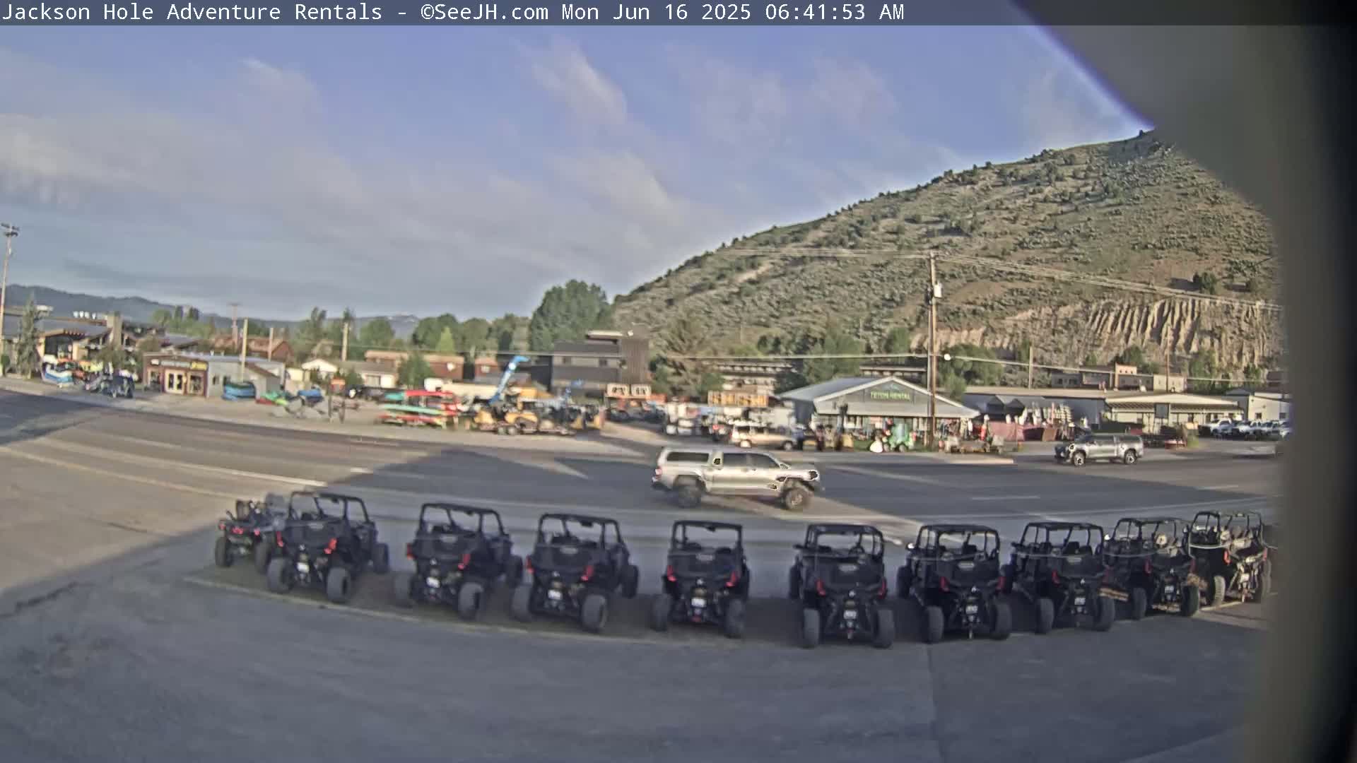 Under a partly cloudy sky, several off-road vehicles are parked in a row in front of a collection of buildings near a hill.