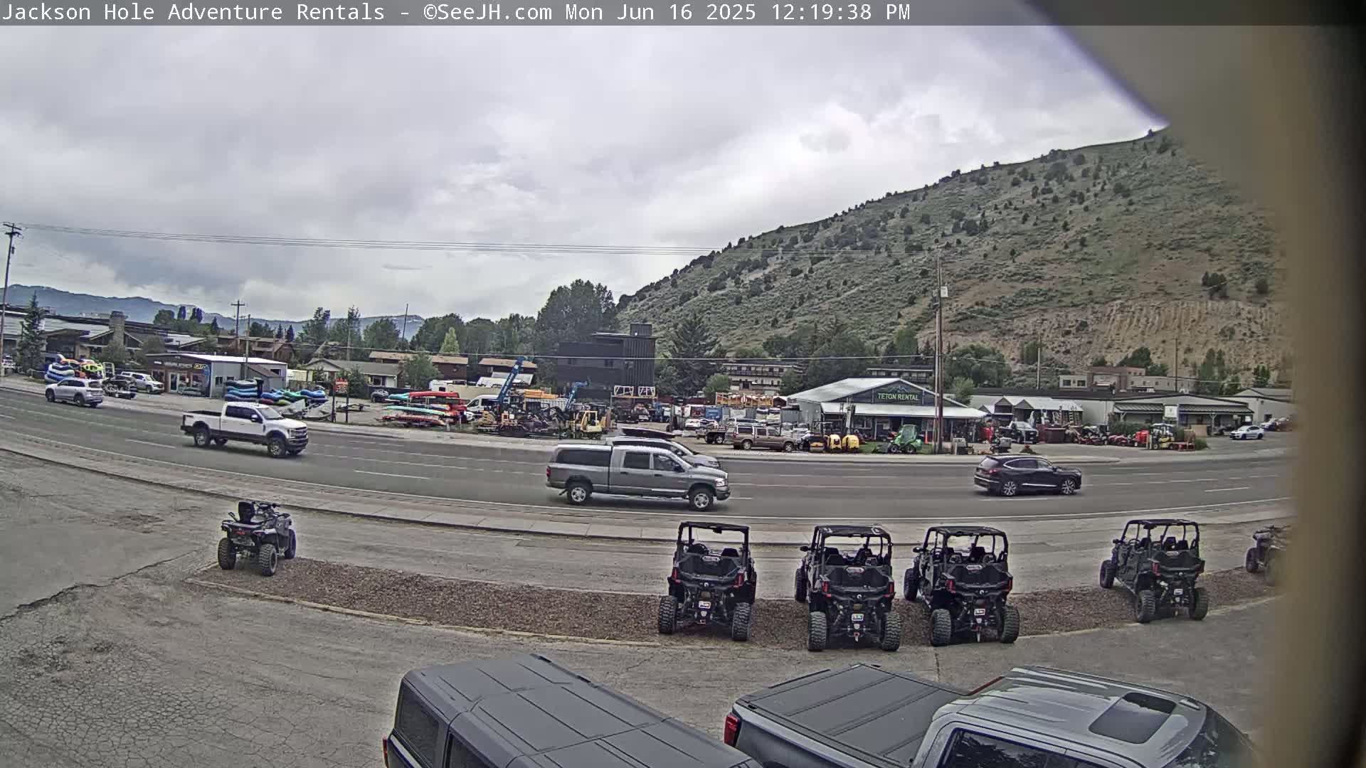 A street scene shows several vehicles parked and driving, with ATVs in the foreground, under an overcast sky in front of a hillside.