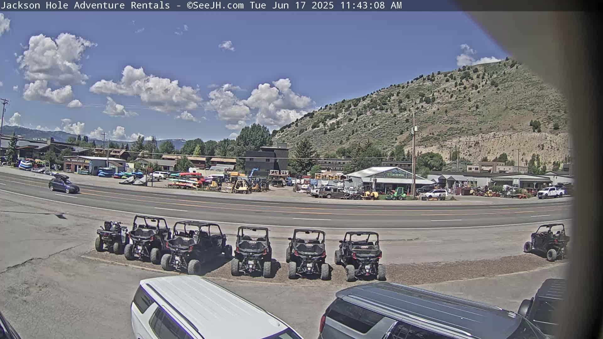 A sunny day with partly cloudy skies shows a parking lot full of all-terrain vehicles in front of a rental business near a mountainside.