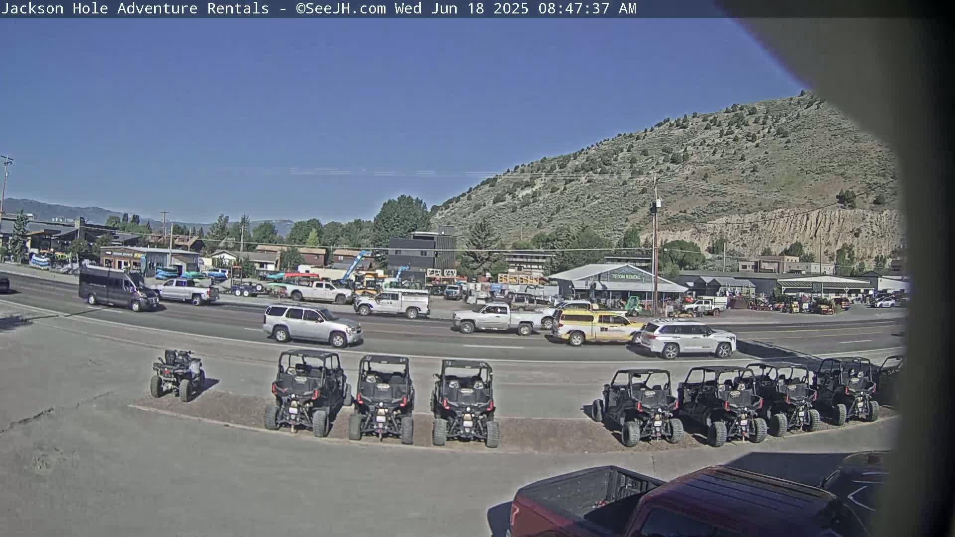 A sunny day reveals a parking lot full of all-terrain vehicles in front of several businesses and a road with cars and trucks driving past a hill.