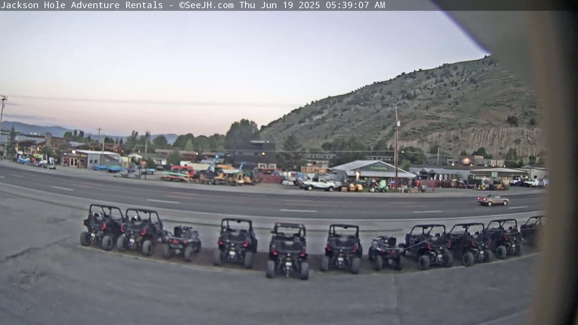 A row of off-road vehicles is parked in a lot across from a street with other vehicles and buildings under a twilight sky.