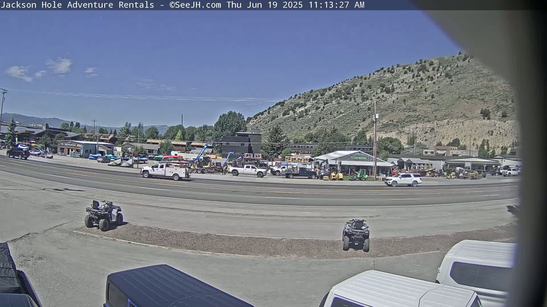 Under a clear blue sky, several vehicles are parked in a lot near a road next to a hill with sparse vegetation.