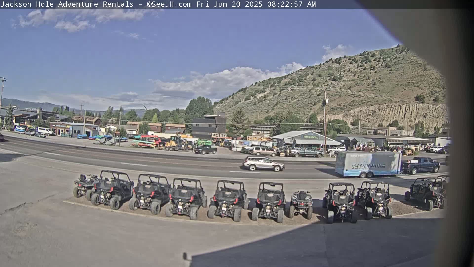 A row of all-terrain vehicles is parked in a lot next to a road, with various businesses and a partly cloudy sky in the background.