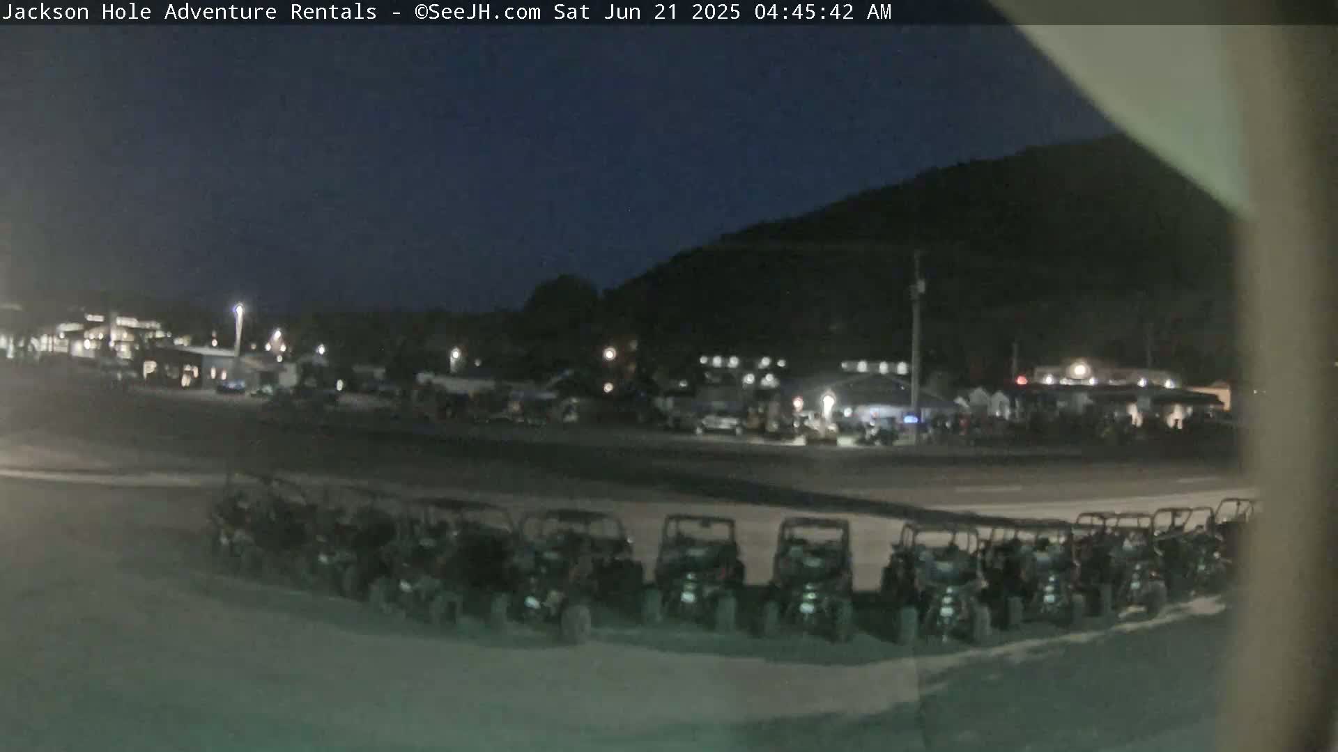 A row of parked off-road vehicles sits in a dark parking lot at night, under a clear, dark sky, with buildings and a hill in the background.