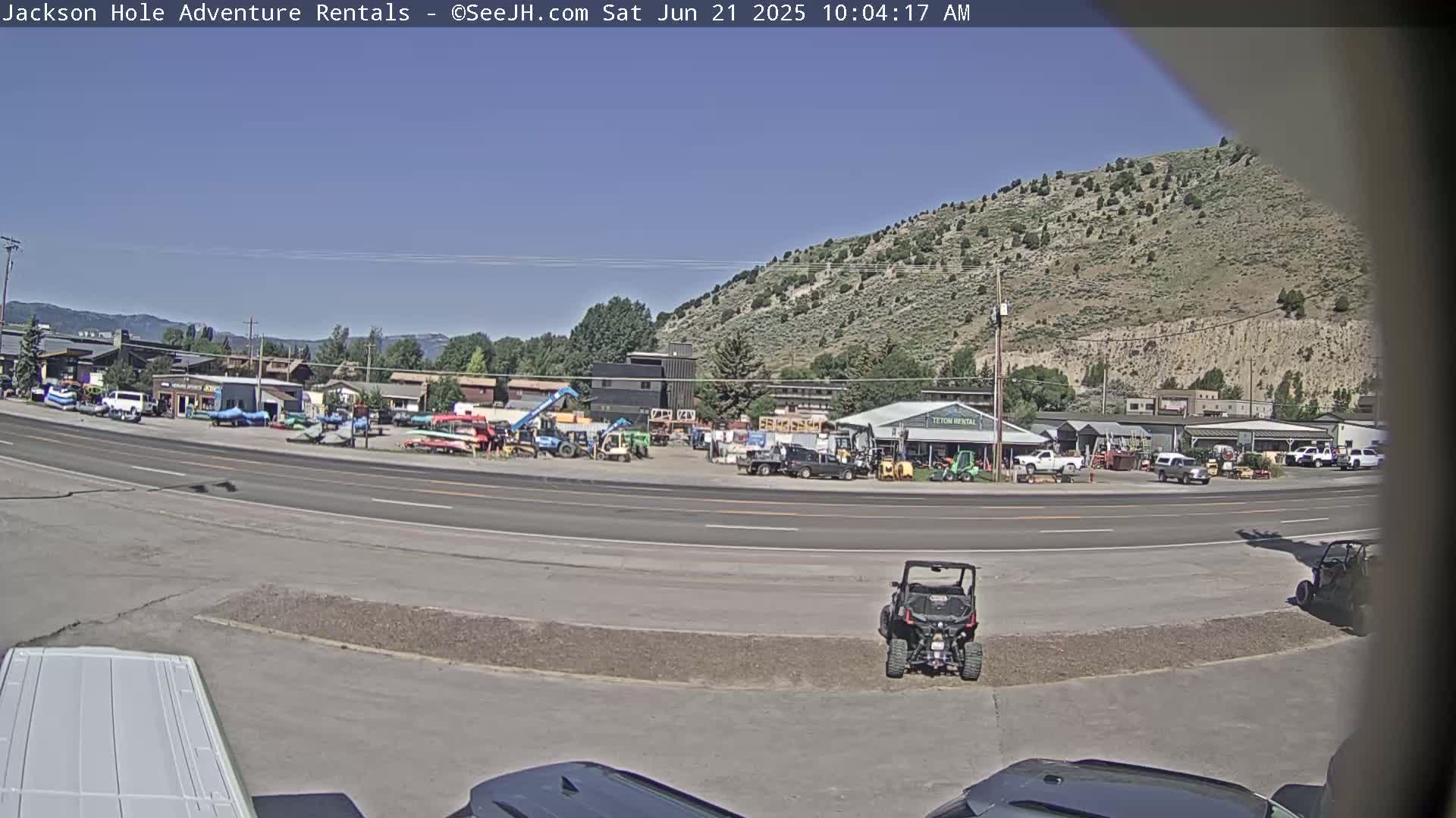 A sunny day reveals a parking lot filled with various vehicles, including ATVs and construction equipment, situated next to a road and a hill with sparse vegetation.
