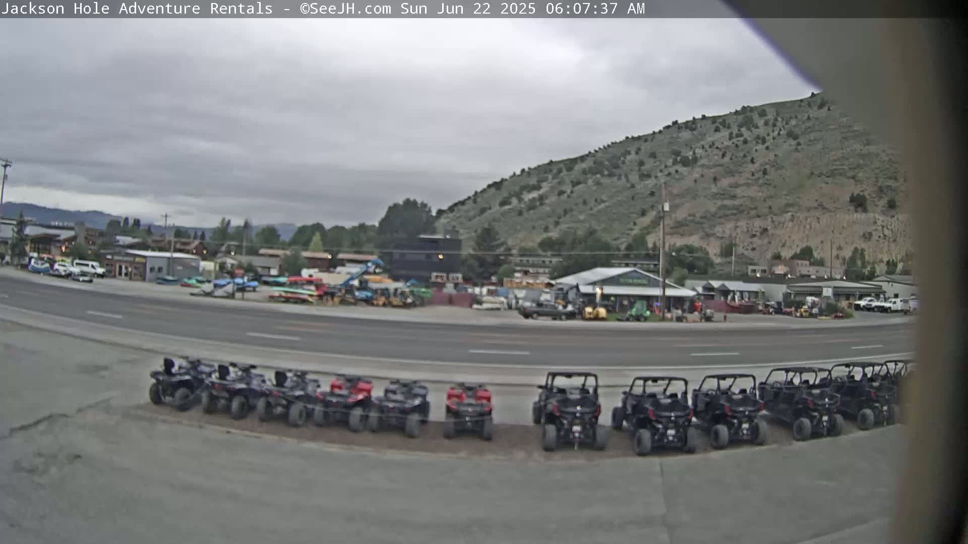 A row of all-terrain vehicles is parked in a lot next to a road in front of several buildings and a hill under an overcast sky.