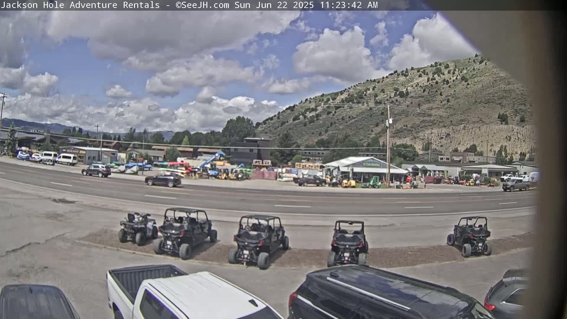 A partly cloudy day reveals a street scene with several ATVs parked in a lot, numerous vehicles in a nearby rental yard, and a mountainside in the background.