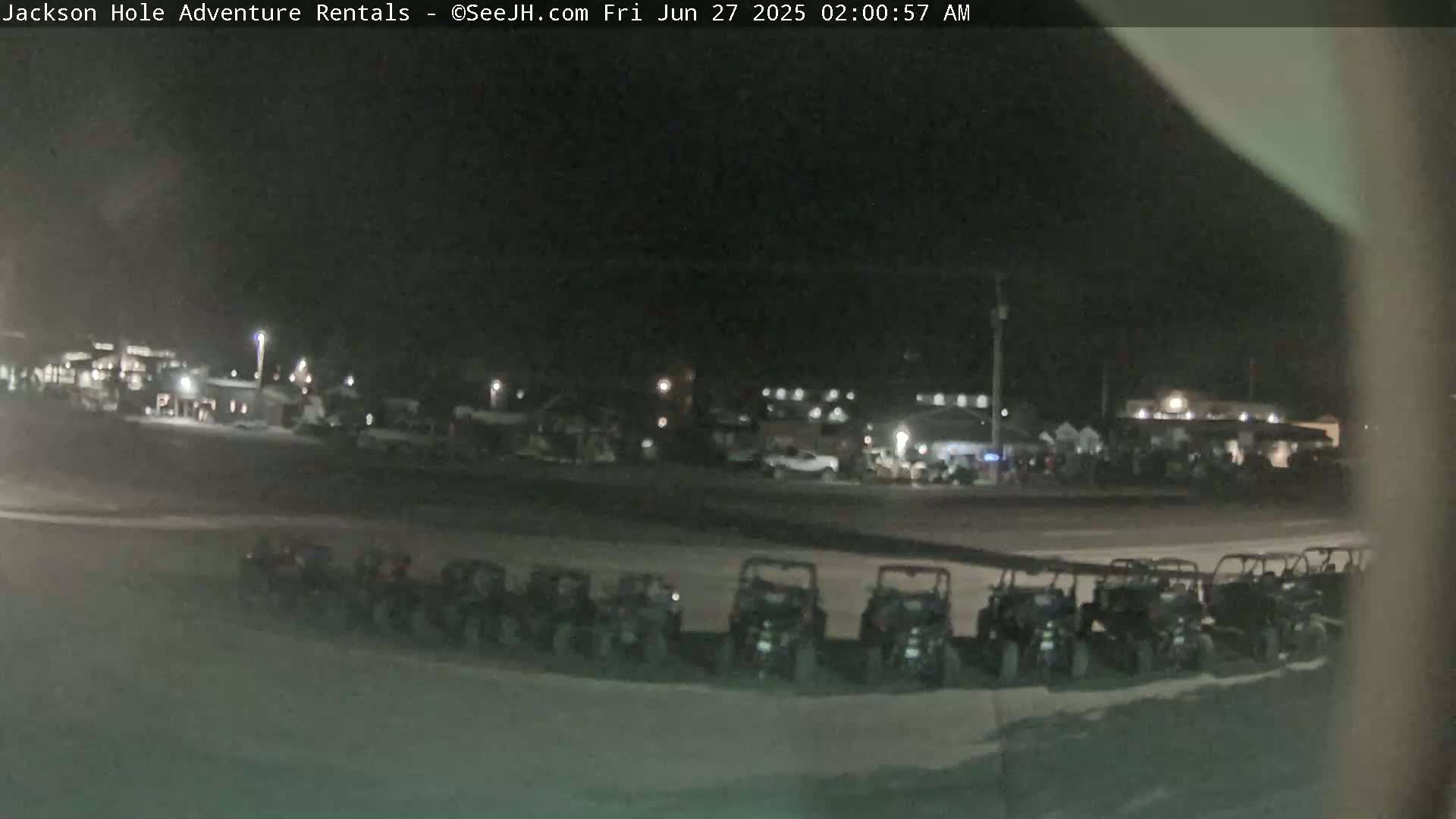 A row of off-road vehicles is parked at night in a mostly empty lot in front of a building with some lights on.