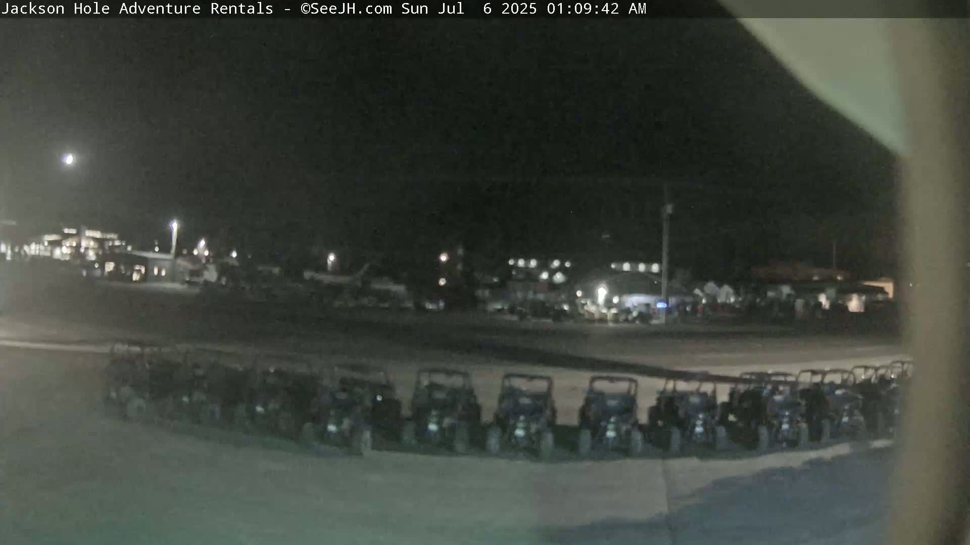 A nighttime, outdoor scene shows a row of parked all-terrain vehicles in a parking lot with other vehicles and buildings visible in the background under a dark sky.