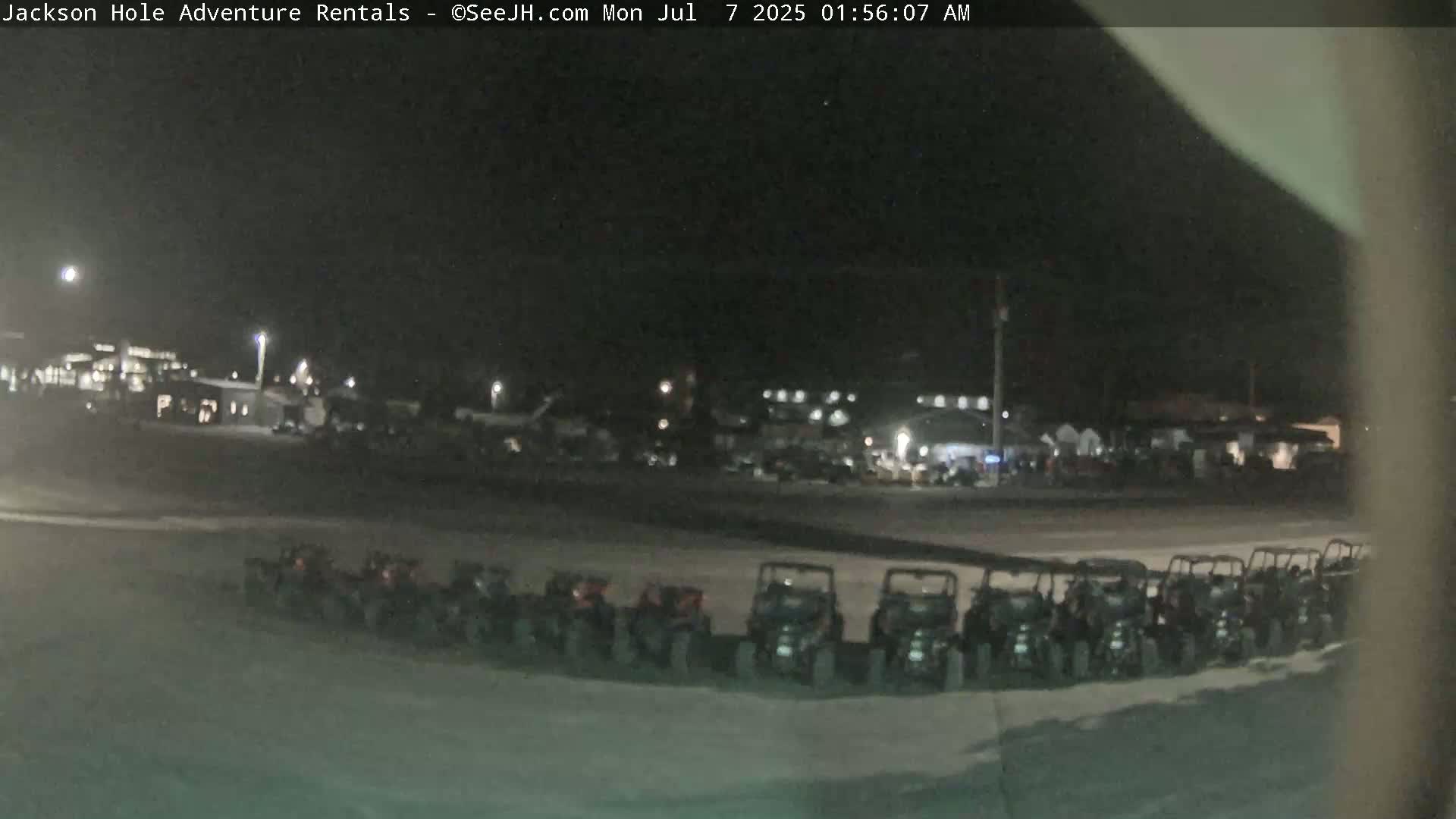 A nighttime, outdoor view shows a row of parked all-terrain vehicles in a parking lot near some buildings.