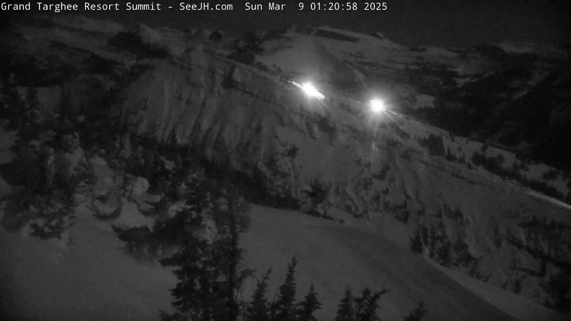 A nighttime, snowy scene shows a sloped, snow-covered landscape with vehicle headlights visible in the distance.