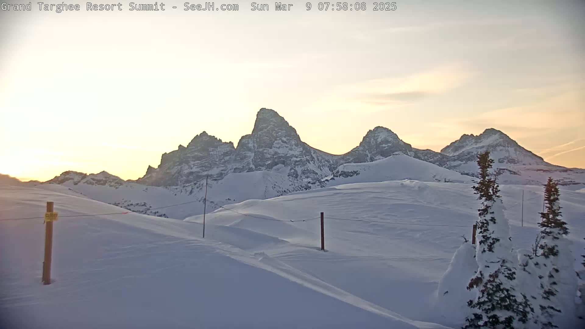 A snow-covered landscape at sunrise shows a range of jagged, snow-capped mountains in the background.