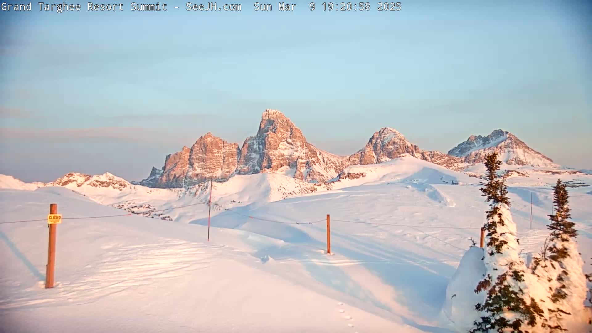 A snow-covered landscape features several prominent, snow-dusted mountain peaks under a clear, light-blue sky at sunrise.