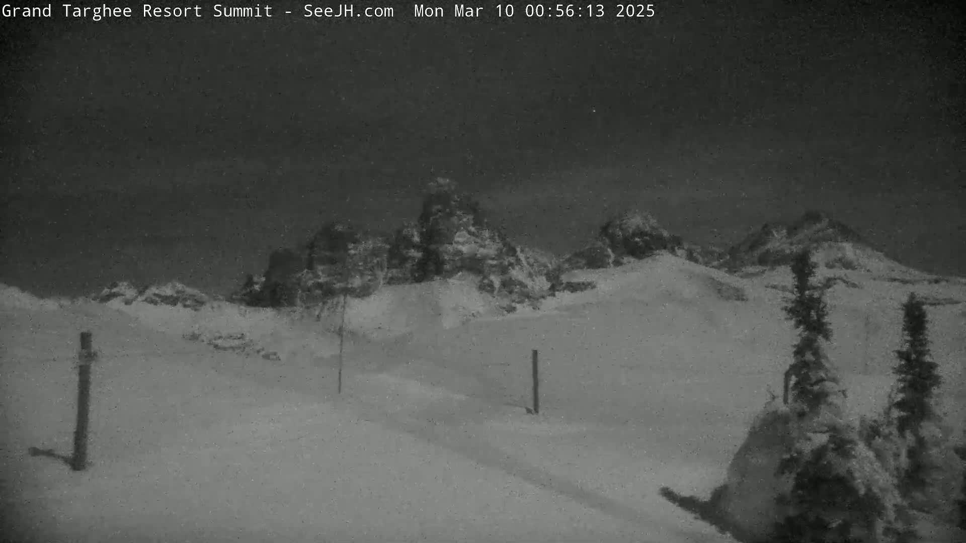 A nighttime, snow-covered landscape shows snow-dusted mountains in the distance and two vertical posts in the foreground under a dark sky.