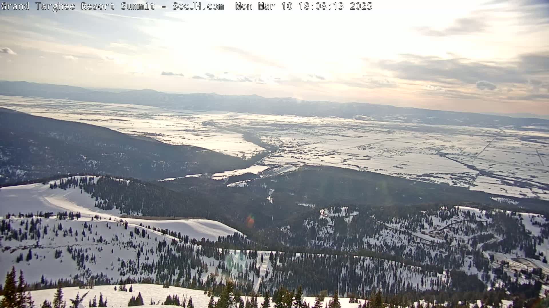 A snow-covered mountain range overlooks a vast, snow-dusted valley under a partly cloudy sky.
