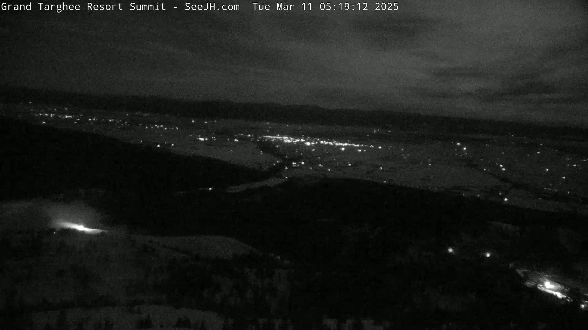A nighttime, high-angle view shows a dark landscape with a distant city illuminated by lights, under a cloudy sky.