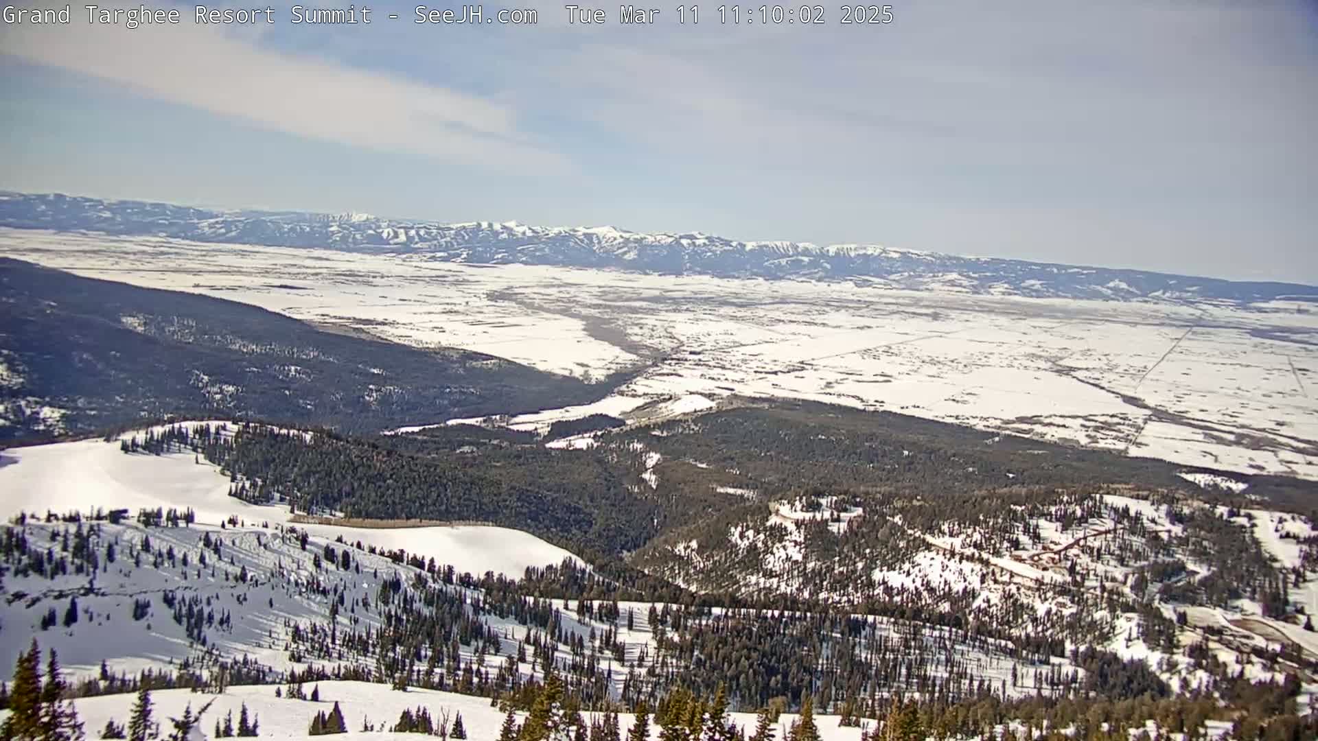 A snow-covered mountain vista stretches to a distant, snow-dusted valley under a mostly clear sky.