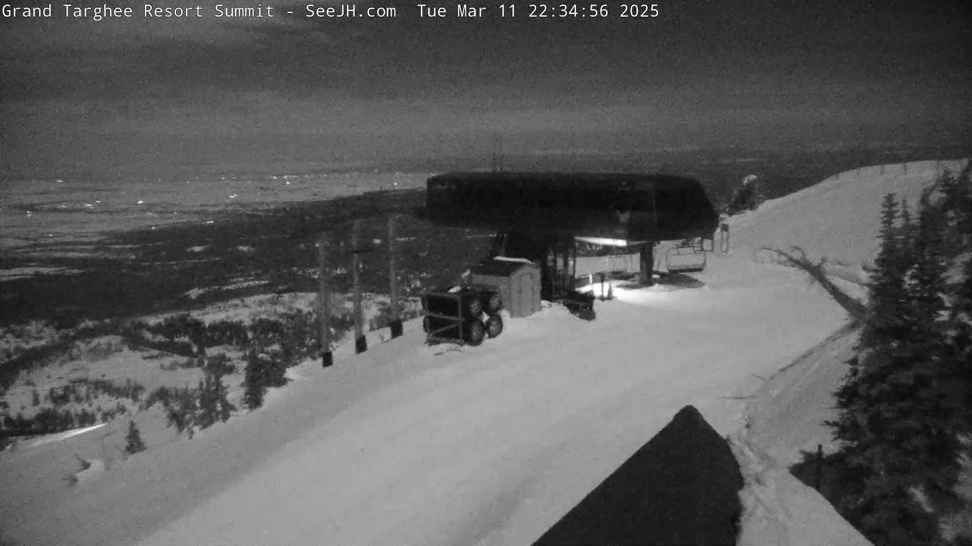 A nighttime view of a snow-covered ski resort at the summit, showing a chairlift and surrounding landscape under a dark sky.