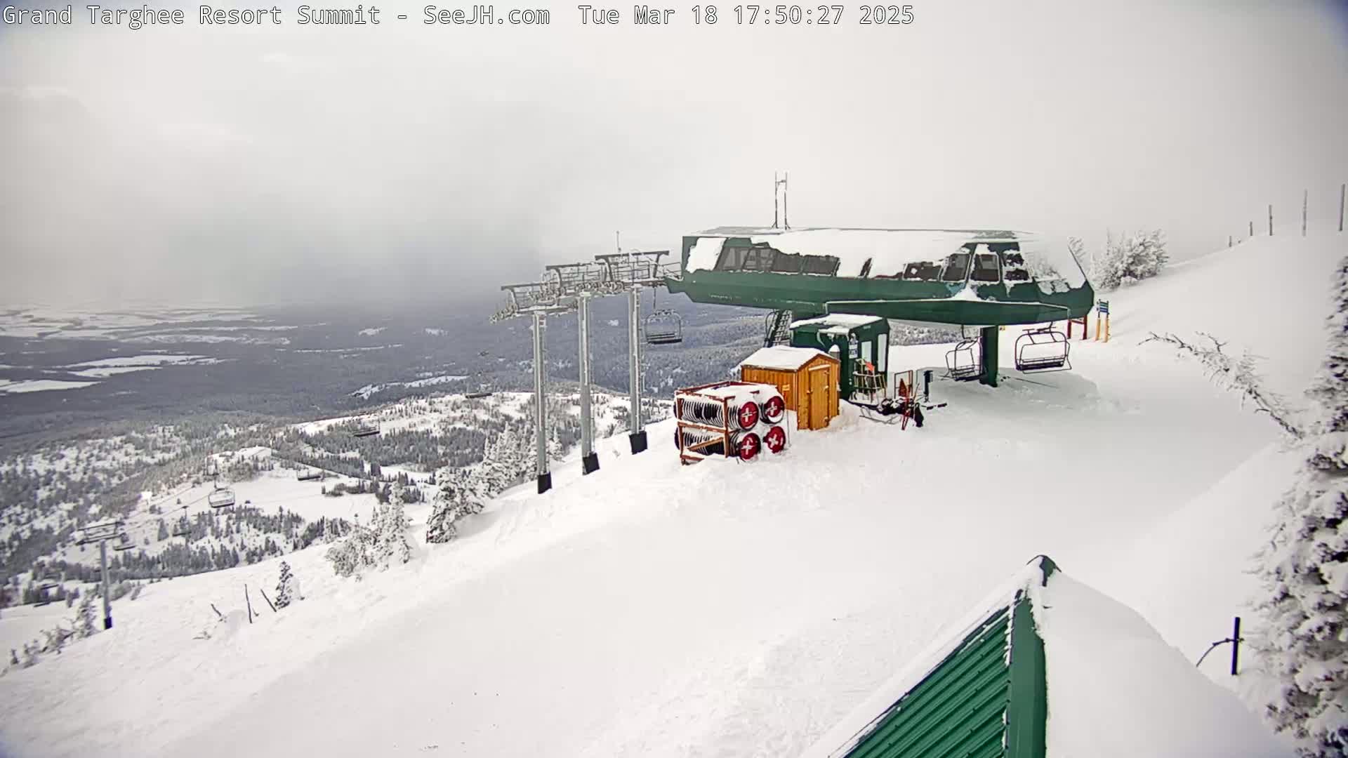 A snow-covered ski lift at a mountain resort overlooks a vast, snow-dusted landscape under an overcast sky.