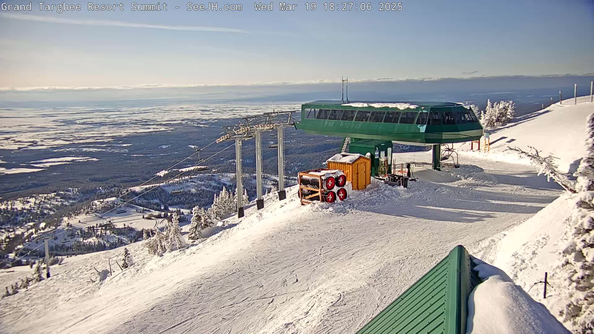 A ski lift at a snowy mountaintop overlooks a vast, snow-covered valley under a clear, sunny sky.