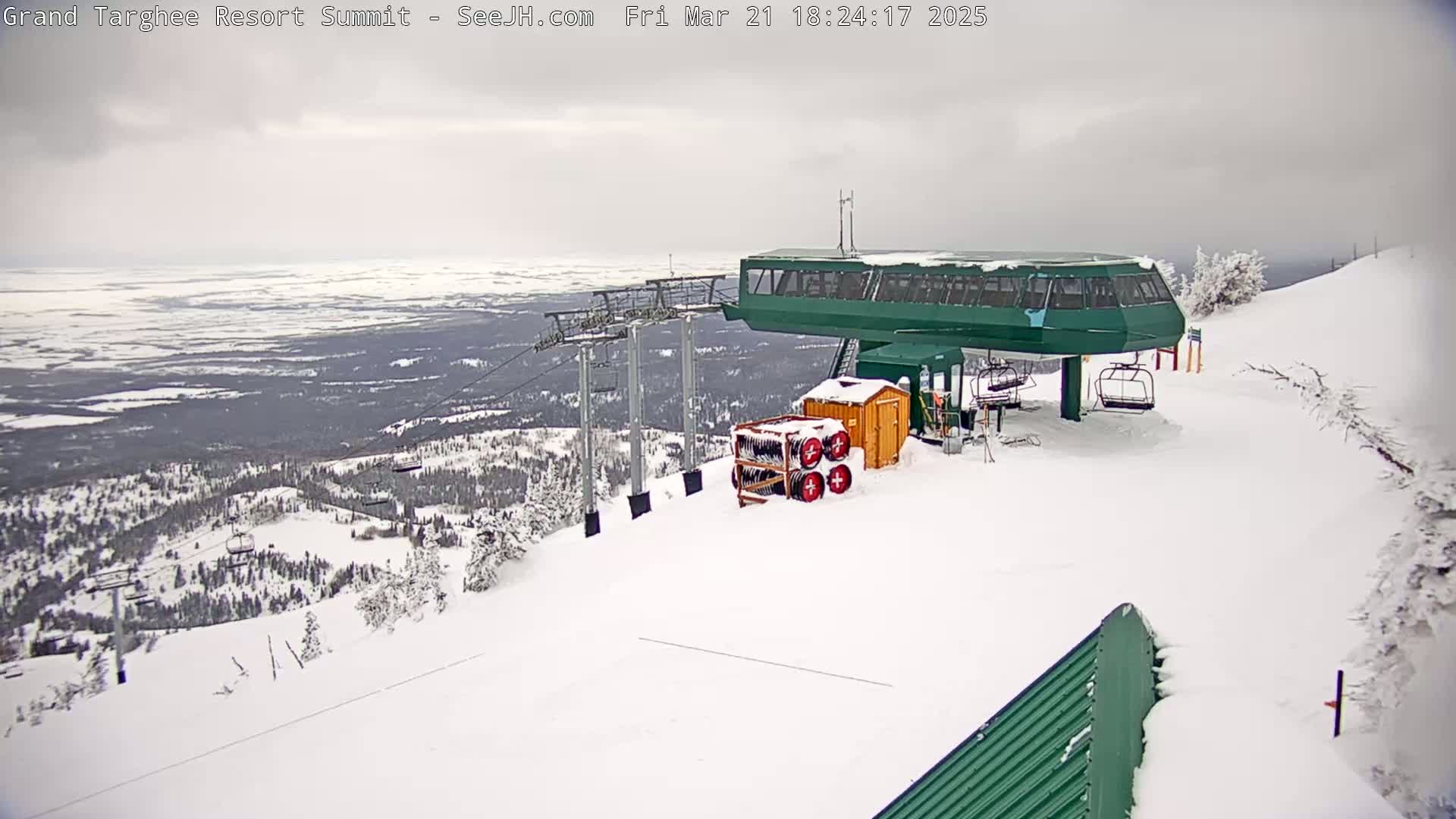 A snow-covered ski resort summit shows a green chairlift station with overcast skies and a view of a vast, snow-dusted landscape.
