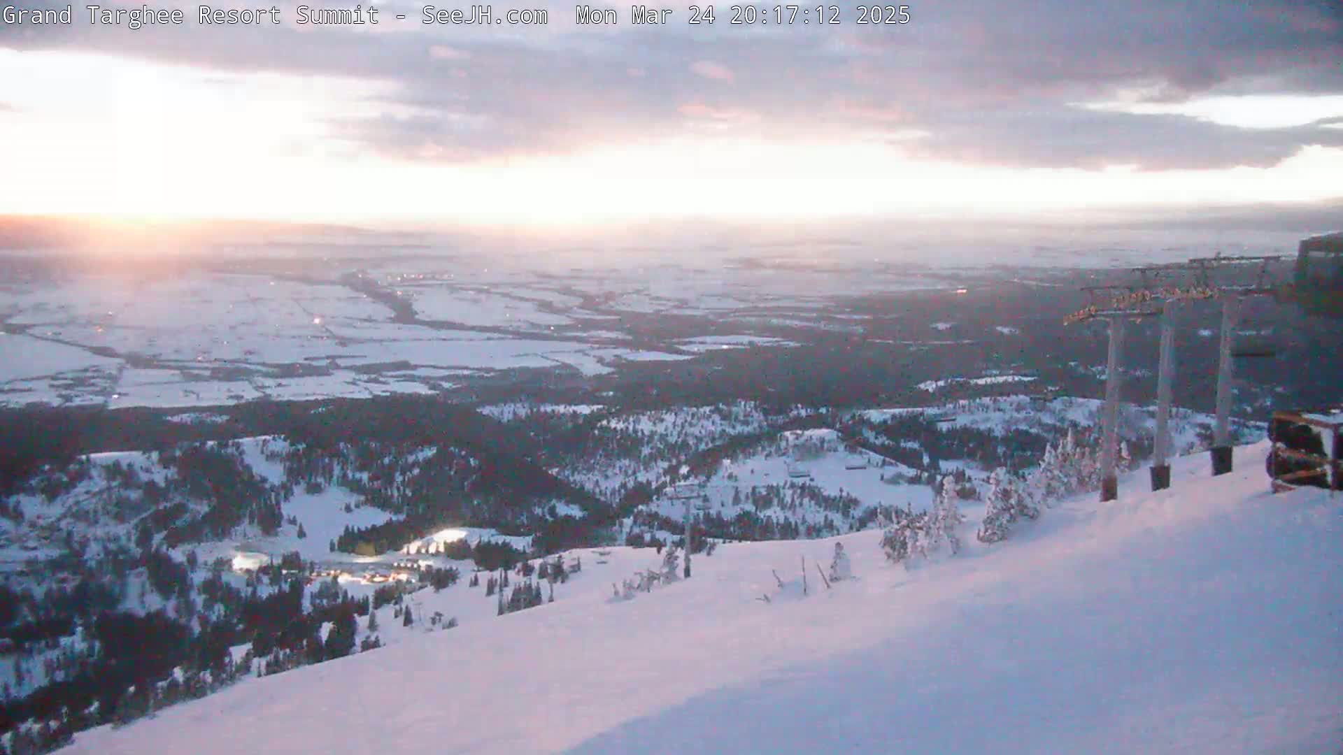 A snow-covered mountain slope at dusk overlooks a valley with a town below, under a partly cloudy sky.