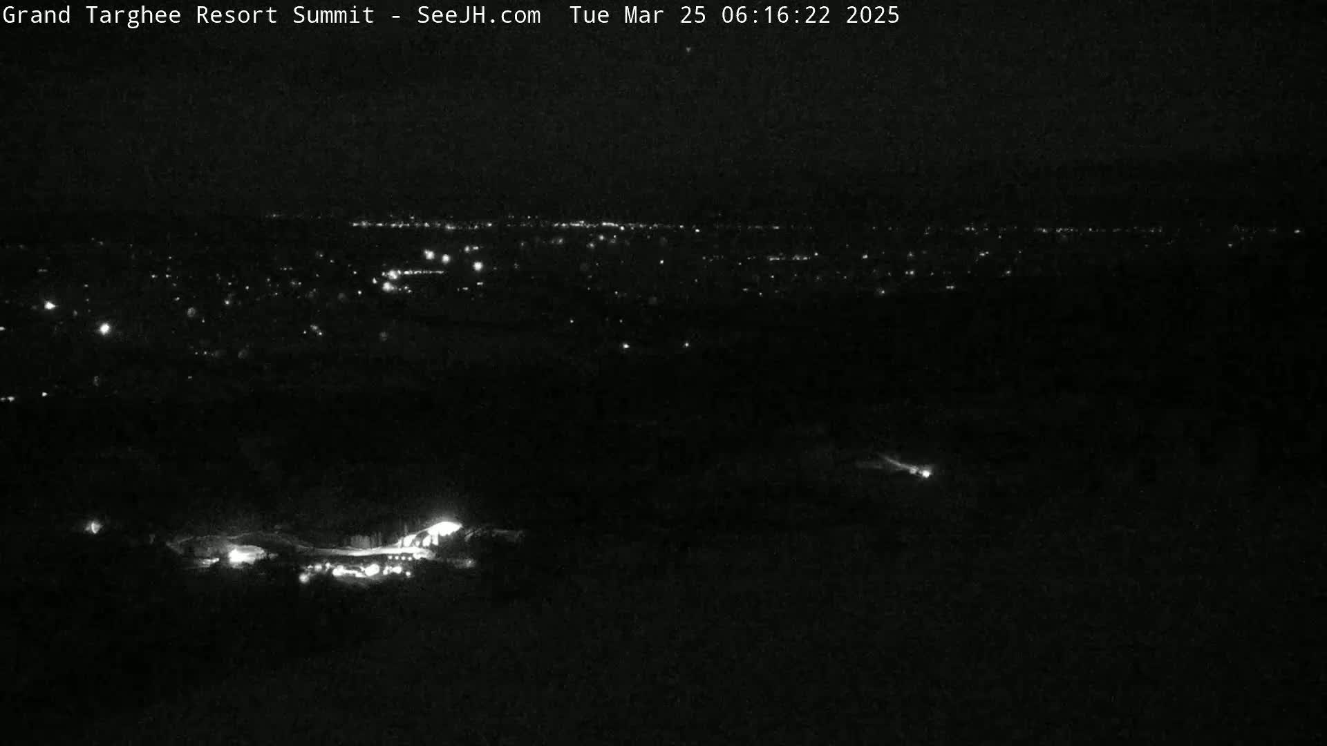 A nighttime, long-range view shows a distant city glowing with scattered lights, while closer to the camera, a few faint lights are visible in the dark landscape.