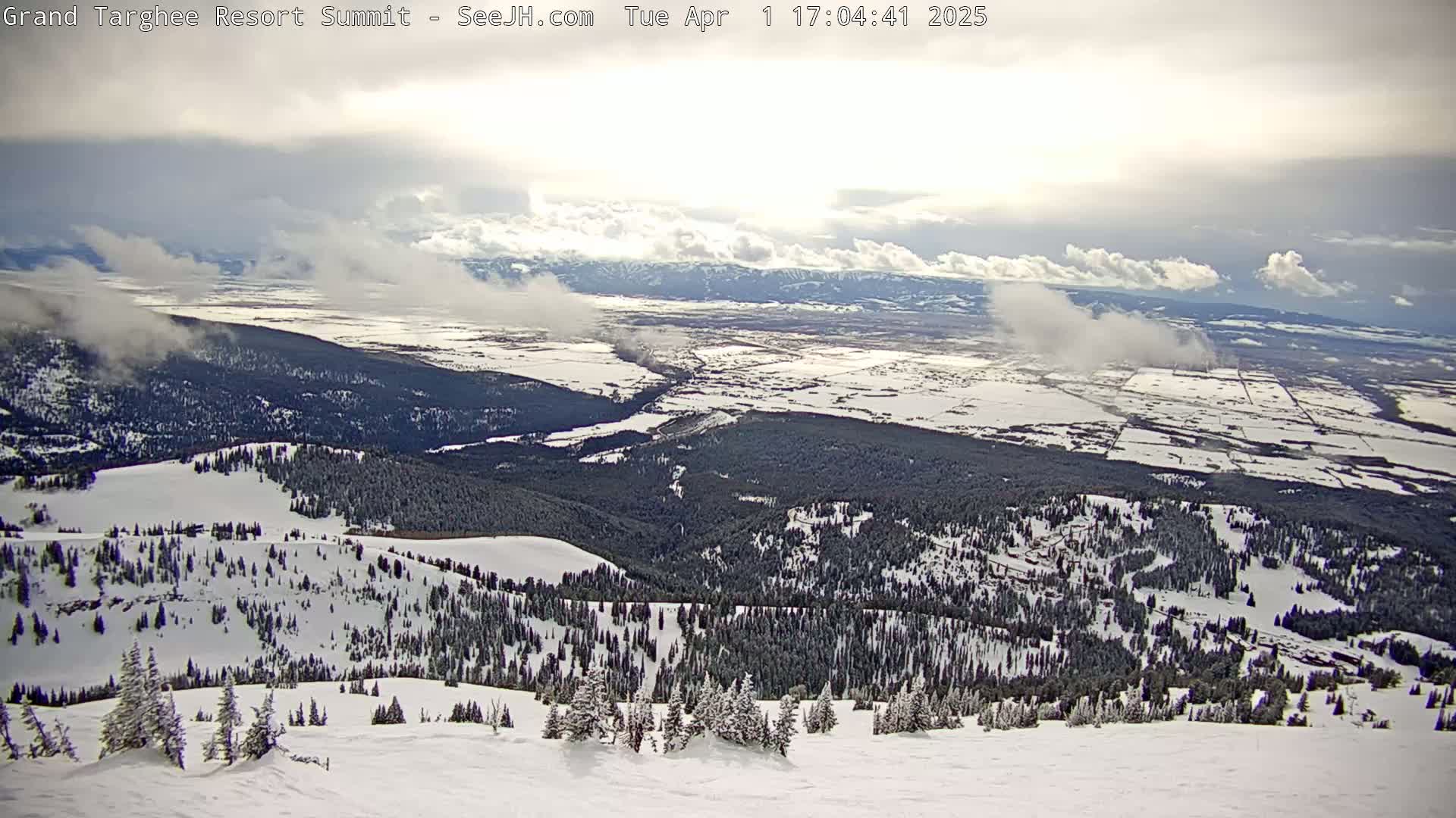 A snow-covered mountain slope overlooks a valley with patchy snow cover under a partly cloudy sky.