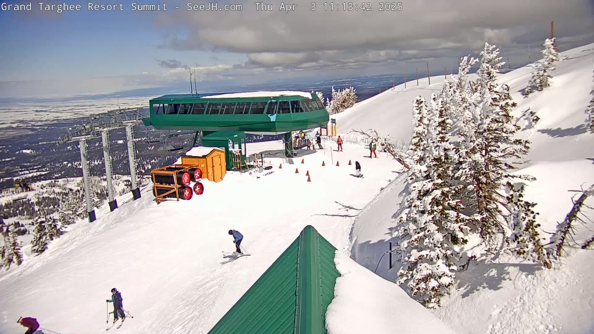 A snow-covered ski resort summit with a green chairlift, several skiers and snowboarders, and evergreen trees dusted with snow under partly cloudy skies.