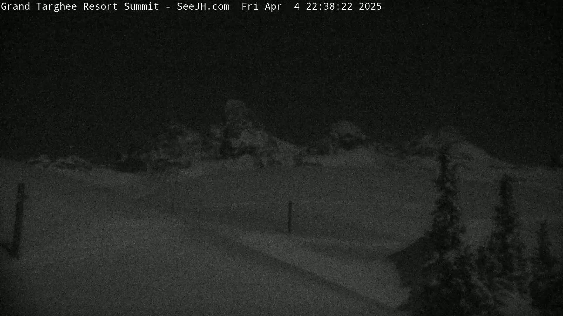 A snow-covered landscape at night shows a road leading towards dimly lit mountains under a dark sky.