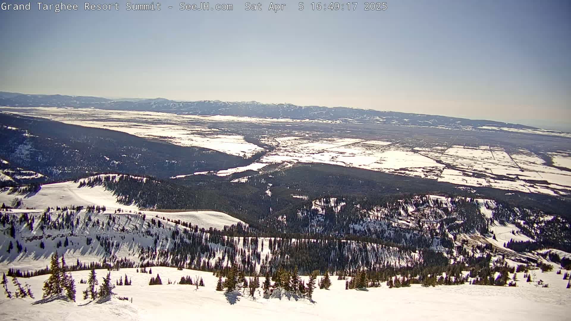 A snow-covered mountaintop reveals a panoramic view of a valley with patches of snow under a clear, bright sky.