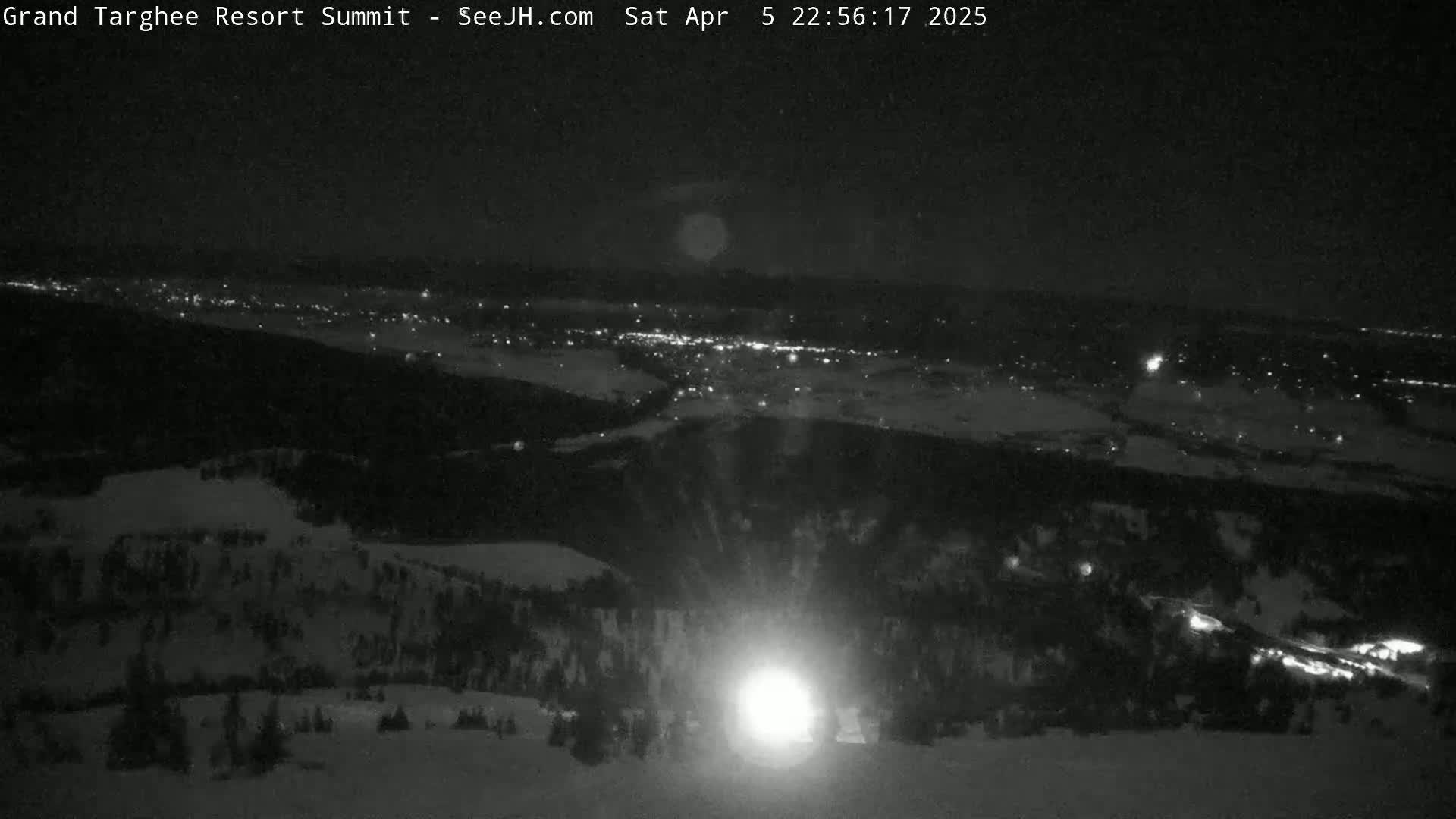 A nighttime, snowy mountaintop view shows a brightly lit area below and a distant town speckled with lights.