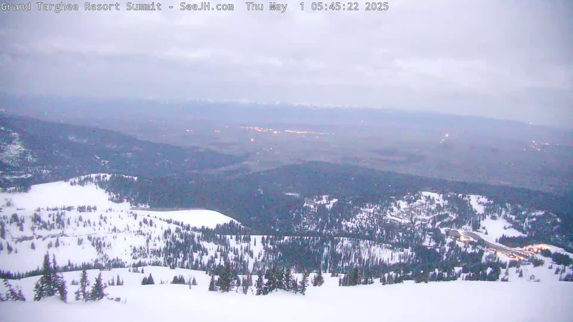 A snow-covered mountain slope overlooks a valley with distant lights under an overcast sky.