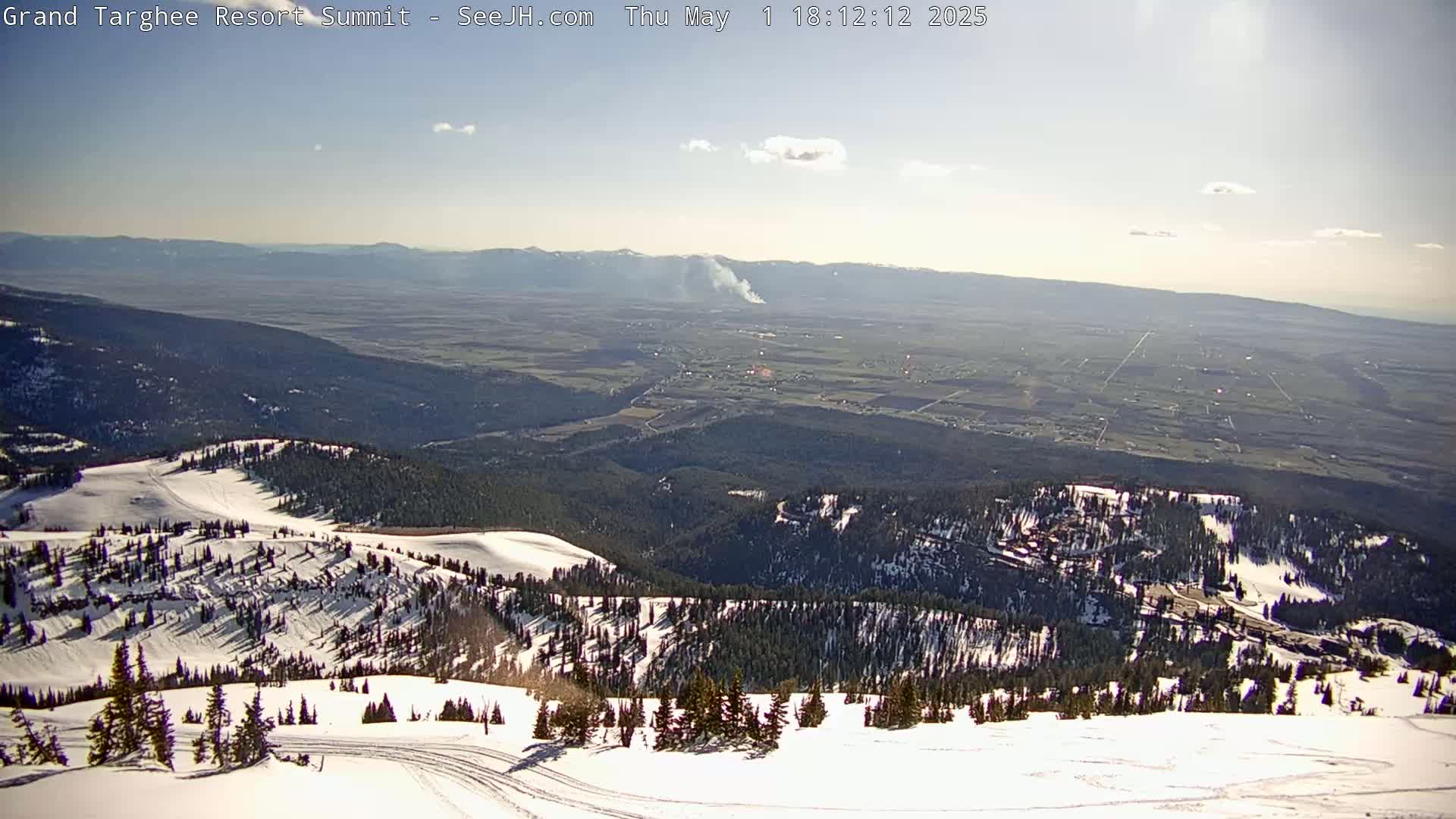 A snow-covered mountaintop offers a panoramic view of a valley with distant mountains under a mostly clear, sunny sky, with a wisp of smoke visible in the distance.