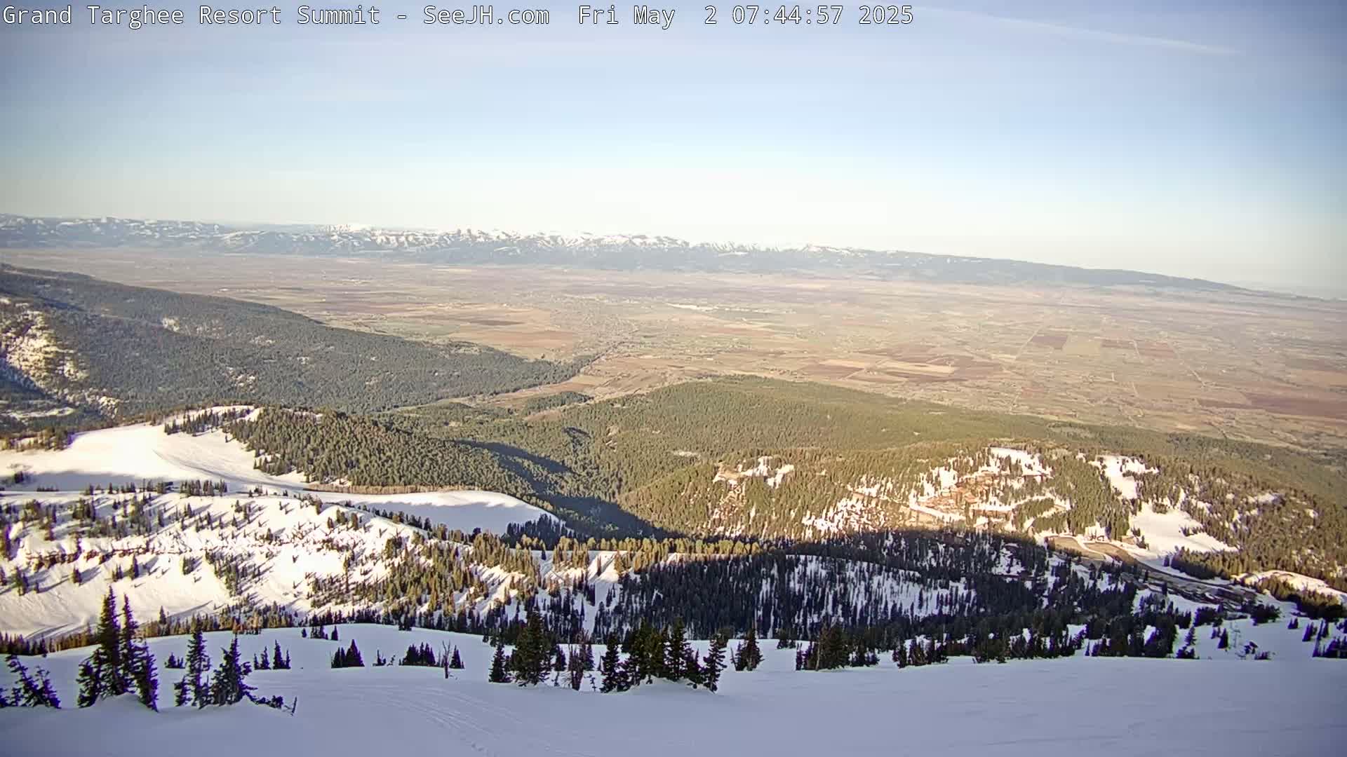 A snowy mountaintop view reveals a vast, flat valley stretching to distant, snow-capped mountains under a clear sky.