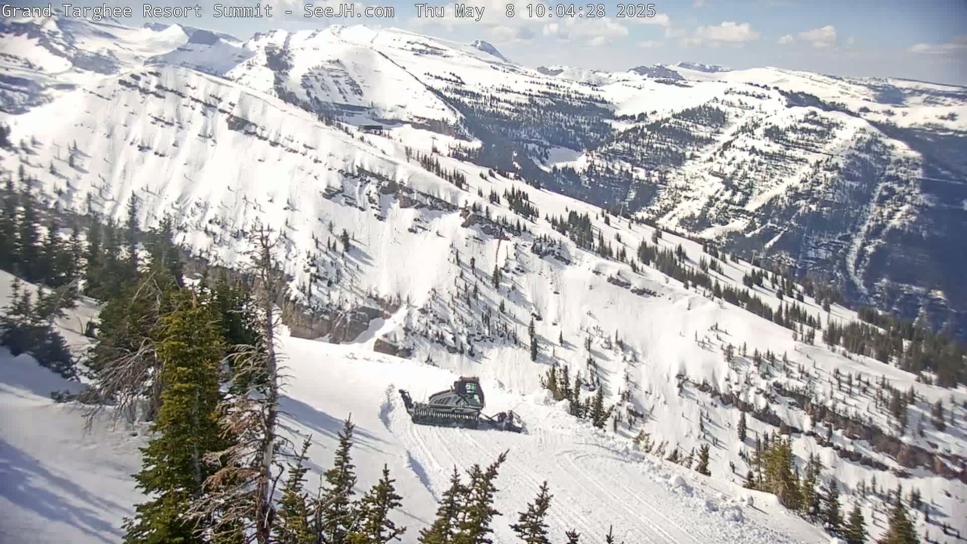 A snowcat grooms a snow-covered mountain slope under a partly sunny sky.