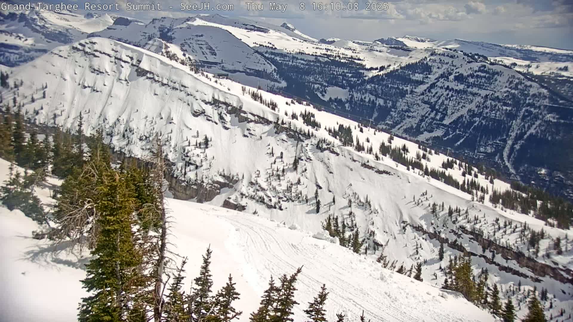 A snow-covered mountain slope with evergreen trees and a partly sunny sky is seen from a high vantage point.
